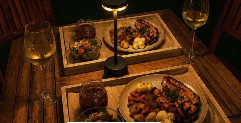 Duas bandejas de madeira na mesa, cada uma com prato principal, salada e taça de vinho branco, sob luminária de mesa